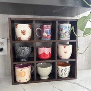 May include: A dark brown wooden display shelf with nine compartments, each holding a different decorative mug or bowl. The mugs and bowls feature various designs, including leaves, hearts, and geometric patterns. The text "Homvex Designs" is visible.