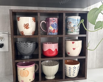May include: A dark brown wooden display shelf with nine compartments, each holding a different decorative mug or bowl. The mugs and bowls feature various designs, including leaves, hearts, and geometric patterns. The text "Homvex Designs" is visible.