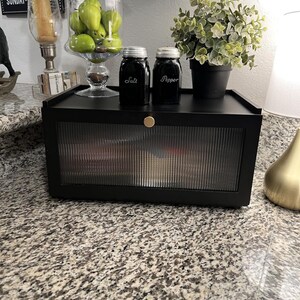 May include: A black cabinet with a ribbed glass door, holding various items. On top, there are two black jars labeled "Salt" and "Pepper", a small potted plant, and a glass jar filled with green fruit. A gold knob is on the cabinet.