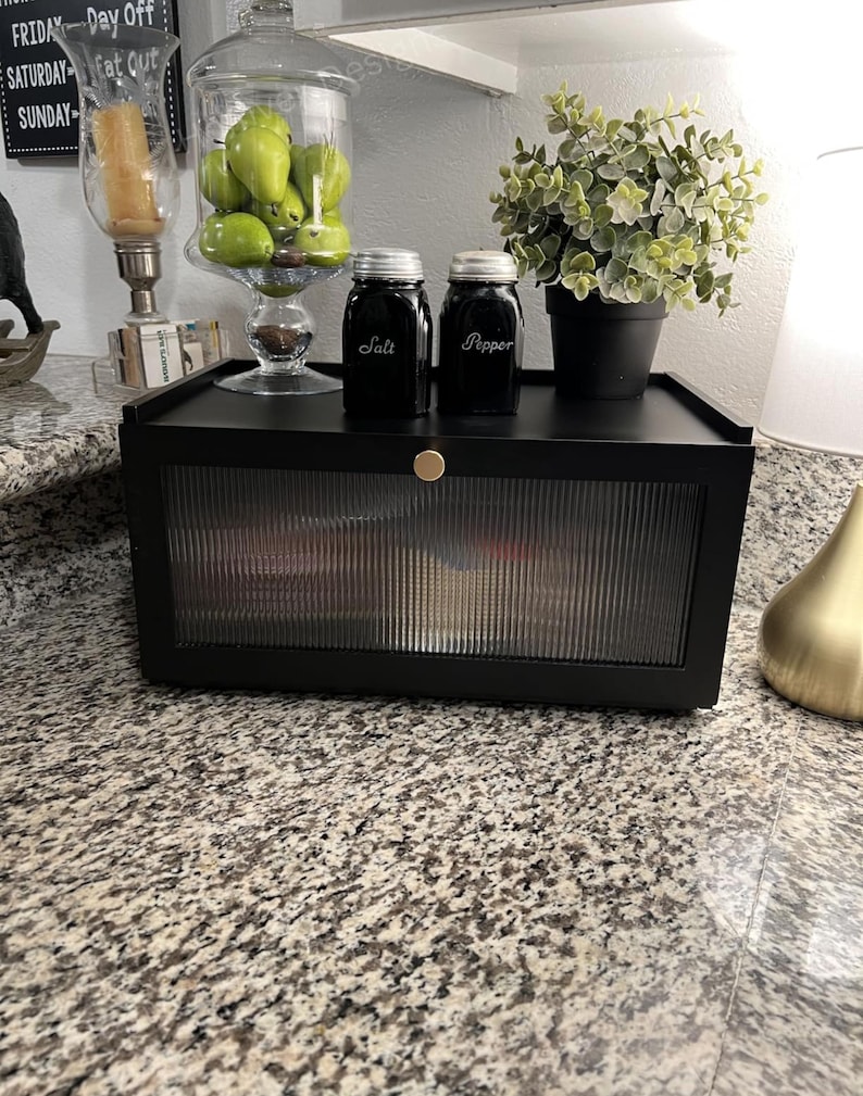 May include: A black cabinet with a ribbed glass door, holding various items. On top, there are two black jars labeled "Salt" and "Pepper", a small potted plant, and a glass jar filled with green fruit. A gold knob is on the cabinet.