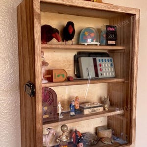 May include: A wooden display case with glass front, filled with miniature vintage items. Includes a radio, snow globe, dreidel, and figurines. The case is made of light brown wood and has a small wooden chest on top.