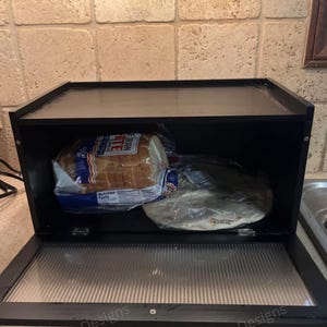 May include: Black bread box with a hinged, clear front door and a clear top. The box contains a loaf of white bread and a package of tortillas. The bread box has a gold-colored handle.