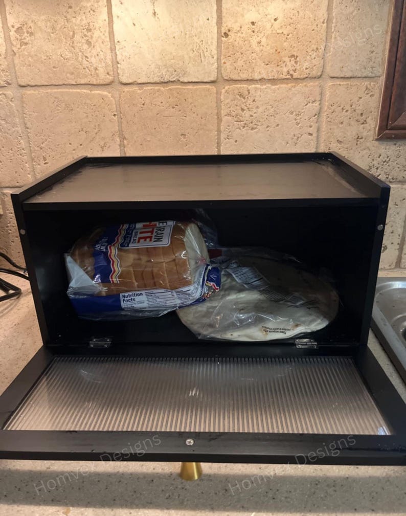 May include: Black bread box with a hinged, clear front door and a clear top. The box contains a loaf of white bread and a package of tortillas. The bread box has a gold-colored handle.