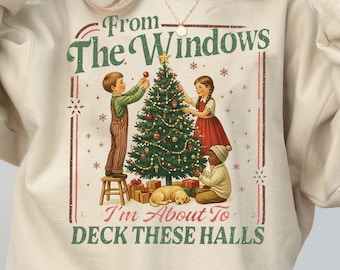 May include: Beige sweatshirt with a festive graphic. The design features a Christmas tree being decorated by three children, with the text "From The Windows I'm About To DECK THESE HALLS". The sweatshirt has long sleeves and a crew neck.