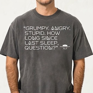 May include: Dark gray t-shirt with the text "GRUMPY. ANGRY. STUPID. HOW LONG SINCE LAST SLEEP, QUESTION?" in white. The shirt has a vintage, worn look.