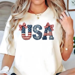 May include: Cream-colored t-shirt featuring the word "USA" in a denim-style font, with the letters filled with the American flag design. Red and white striped bows accent the top of the letters. A patriotic themed tee.