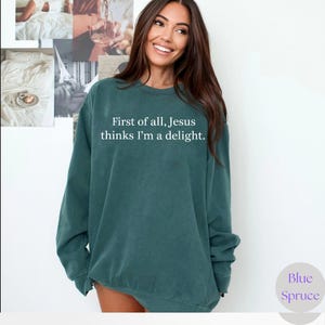 May include: Teal-colored sweatshirt with the text "First of all, Jesus thinks I'm a delight." The sweatshirt has long sleeves and a crew neck. The brand name "Blue Spruce" is in a circle on the bottom right.