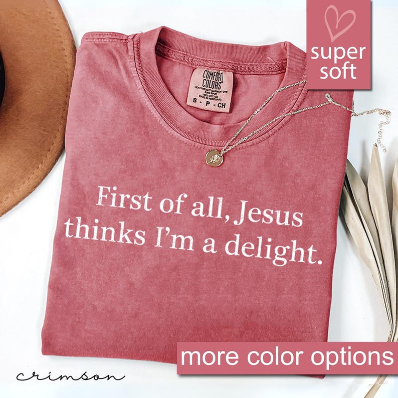 May include: Crimson-colored t-shirt with the text "First of all, Jesus thinks I'm a delight." A gold pendant necklace rests on the shirt. The shirt has a "super soft" label and a "more color options" label.