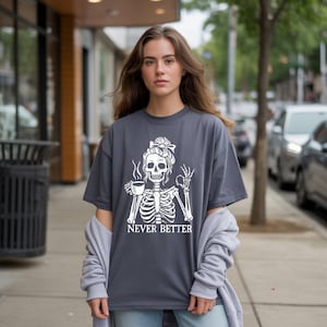 May include: A dark gray t-shirt featuring a white skeleton design. The skeleton is holding a coffee cup and making an "okay" hand gesture. The text "NEVER BETTER" is printed below the skeleton. The shirt is a casual, oversized fit.