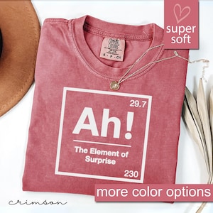 May include: Crimson-colored t-shirt with the text "Ah! The Element of Surprise" in a white square. A silver necklace with a pendant rests on the shirt. The shirt has a crew neck and short sleeves.