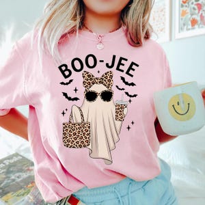 May include: Pink t-shirt with the text "BOO-JEE" above a cartoon ghost wearing sunglasses and a leopard print bow, holding a leopard print tote bag and a coffee cup. Black bats and stars surround the ghost. A white mug with a yellow smiley face is in the background.