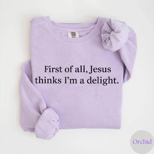 May include: A light purple sweatshirt with the text "First of all, Jesus thinks I'm a delight." The sweatshirt is made of a soft material and has a crew neck. The sleeves are folded. The brand name "Comfort Colors" is visible on the tag.