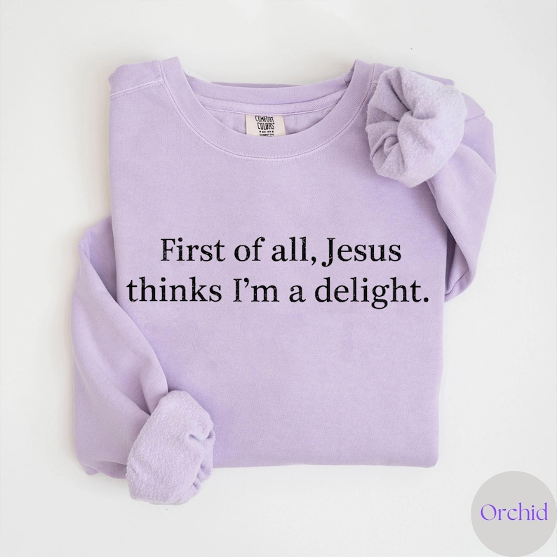 May include: A light purple sweatshirt with the text "First of all, Jesus thinks I'm a delight." The sweatshirt is made of a soft material and has a crew neck. The sleeves are folded. The brand name "Comfort Colors" is visible on the tag.