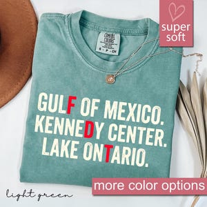 以下が含まれることがあります： クルーネックとComfort Colorsのブランドラベルが付いたライトグリーンのTシャツ。シャツには「GULF OF MEXICO. KENNEDY CENTER. LAKE ONTARIO.」という白い文字がプリントされており、F、D、Tの文字が赤色で強調されています。シルバーのネックレスがシャツの上に置かれています。