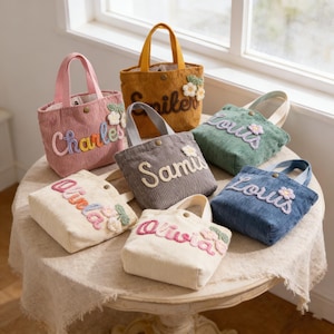 Puede incluir: Una colección de pequeños bolsos de mano de pana en varios colores, cada uno bordado con un nombre y adornos florales. Los bolsos tienen asas cortas y un cierre de botón. Los colores incluyen rosa, mostaza, verde, azul, gris y crema.