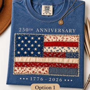 May include: A blue Comfort Colors t-shirt with a sewn-on American flag design. The flag is made of various patterned fabrics and features the text "250th Anniversary" and "1776-2026". A gold necklace with a compass pendant rests on the shirt.