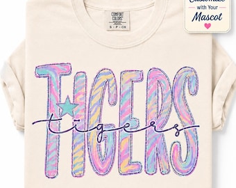 May include: A cream-colored t-shirt featuring the word TIGERS in large, pastel-colored, zebra-striped block letters with a teal star accent. A dark purple cursive script overlays the word. A tag reads Customize with Your Mascot.