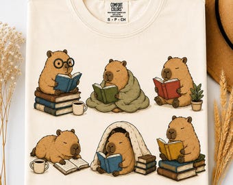 May include: A cream-colored t-shirt featuring a graphic print of six brown capybaras in various reading poses. Some capybaras are sitting on stacks of books, one is wrapped in a blanket, and another is reading under a blanket tent. The shirt has a Comfort Colors label.