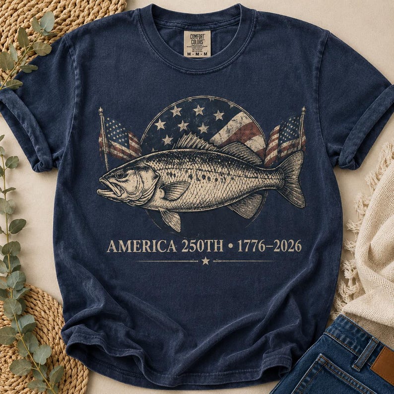 May include: A navy blue short-sleeve t-shirt featuring a graphic of a fish centred in front of a circular American flag design. Below the fish, the text reads AMERICA 250TH &bull; 1776-2026. The shirt has a crew neckline and a Comfort Colors brand label.