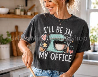 May include: A charcoal gray t-shirt featuring a graphic of a light blue stand mixer, a whisk, a cookie, and hearts. The text reads I'LL BE IN MY OFFICE in white capital letters. The design includes small colorful stars and dots as accents.