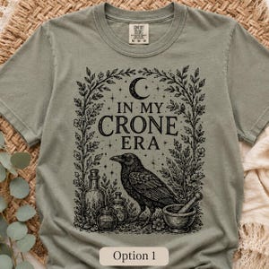 K&ouml;nnte beinhalten: Ein salbeigr&uuml;nes Kurzarm-T-Shirt mit einem schwarzen Grafikdruck, der einen Raben, einen M&ouml;rser mit St&ouml;&szlig;el und Glasflaschen zwischen Ranken zeigt. Unter einer Mondsichel und Sternen steht der Text IN MY CRONE ERA auf dem T-Shirt.