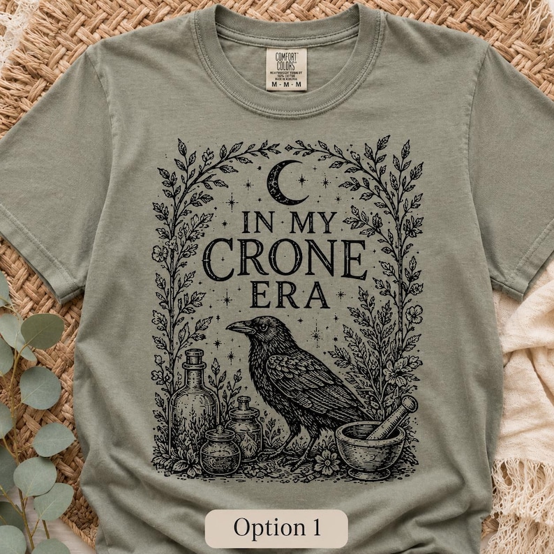 K&ouml;nnte beinhalten: Ein salbeigr&uuml;nes Kurzarm-T-Shirt mit einem schwarzen Grafikdruck, der einen Raben, einen M&ouml;rser mit St&ouml;&szlig;el und Glasflaschen zwischen Ranken zeigt. Unter einer Mondsichel und Sternen steht der Text IN MY CRONE ERA auf dem T-Shirt.