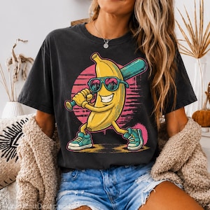 May include: Black t-shirt featuring a cartoon banana wearing sunglasses and holding a baseball bat. The banana is yellow with blue shoes and a pink background. The shirt is worn with blue jean shorts.
