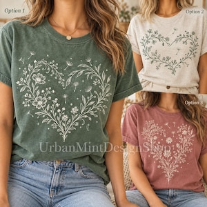 Könnte beinhalten: Ein kurzärmeliges T-Shirt mit Rundhalsausschnitt und einem großen herzförmigen Motiv aus zarten Blumenranken, Blättern und kleinen Blüten. Das Design ist auf drei verschiedenfarbigen T-Shirts abgebildet: Dunkelgrün, Hellgrau und gedämpftes Rot.