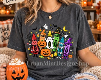 May include: A charcoal gray t-shirt featuring a graphic of colorful crayons dressed in Halloween costumes, including a ghost, a mummy, and a witch. The design includes pumpkins, bats, spiders, and candy corn. The text UrbanMintDesignShop is printed below.