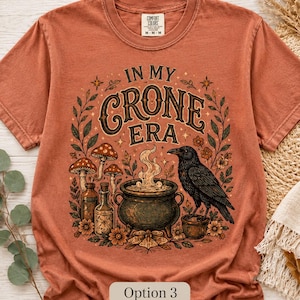 K&ouml;nnte beinhalten: Ein rostfarbenes Kurzarm-T-Shirt mit einem Grafikdruck und dem Text IN MY CRONE ERA. Das Design zeigt einen schwarzen Raben, einen dampfenden Kessel, Glasflaschen, Pilze und florale Elemente in Braun-, Gr&uuml;n- und Oranget&ouml;nen.
