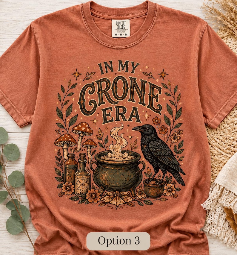 K&ouml;nnte beinhalten: Ein rostfarbenes Kurzarm-T-Shirt mit einem Grafikdruck und dem Text IN MY CRONE ERA. Das Design zeigt einen schwarzen Raben, einen dampfenden Kessel, Glasflaschen, Pilze und florale Elemente in Braun-, Gr&uuml;n- und Oranget&ouml;nen.