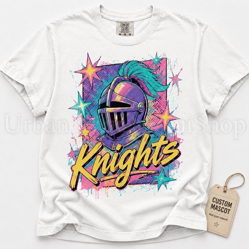 May include: A white short-sleeve t-shirt featuring a graphic of a purple knight helmet with a teal plume. The design includes yellow text reading Knights, surrounded by colorful stars and paint splatters. A tan tag labeled Custom Mascot is attached.