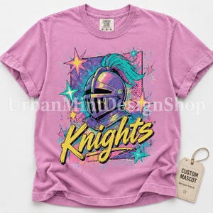 May include: A mauve short-sleeve t-shirt featuring a graphic of a knight helmet with a teal plume, surrounded by colorful stars and the word Knights in yellow script. A tag attached to the shirt reads CUSTOM MASCOT.