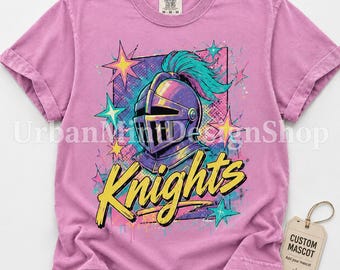 May include: A mauve short-sleeve t-shirt featuring a graphic of a knight helmet with a teal plume, surrounded by colorful stars and the word Knights in yellow script. A tag attached to the shirt reads CUSTOM MASCOT.