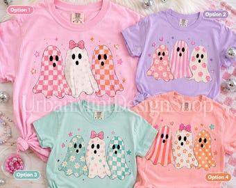 May include: Four t-shirts in pink, lavender, mint green, and peach, each featuring a graphic of three ghosts. The ghosts are decorated with patterns including checkers, flowers, hearts, and smiley faces, with some wearing small pink bows.