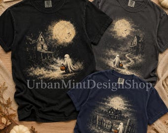 May include: Three short-sleeve t-shirts in black, charcoal, and navy blue, each featuring a graphic print of a ghost holding a pumpkin in front of a house under a full moon. The shirts have a tag that reads "Want custom text or image? Message us!"