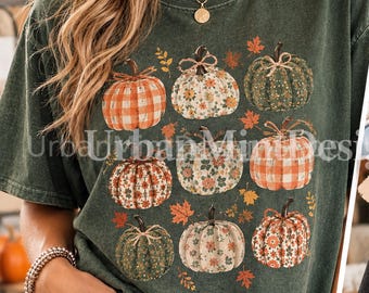May include: A dark green short-sleeve t-shirt featuring a graphic print of nine pumpkins in various patterns, including orange plaid, floral, and solid green, interspersed with small orange and brown autumn leaves and the text UrbanMintDesigns.