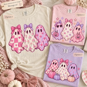 May include: Three t-shirts in cream, light pink, and light purple featuring graphic prints of ghosts with bows, hearts, and pumpkins. The cream shirt shows three ghosts with checkered, floral, and heart patterns. The pink and purple shirts display ghosts with accessories and seasonal motifs.