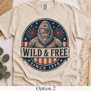Könnte beinhalten: Cremefarbenes T-Shirt mit einem Bigfoot-Motiv, das eine Sonnenbrille im Stil der amerikanischen Flagge trägt. Das Design enthält den Text "WILD & FREE SINCE 1776" in einem kreisförmigen Emblem. Marke Comfort Colors.