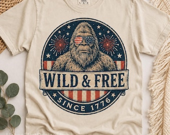 May include: A cream-coloured t-shirt with a Bigfoot graphic wearing sunglasses in the style of the American flag. The design includes the text "WILD & FREE SINCE 1776" within a circular emblem. The shirt is a Comfort Colors brand.