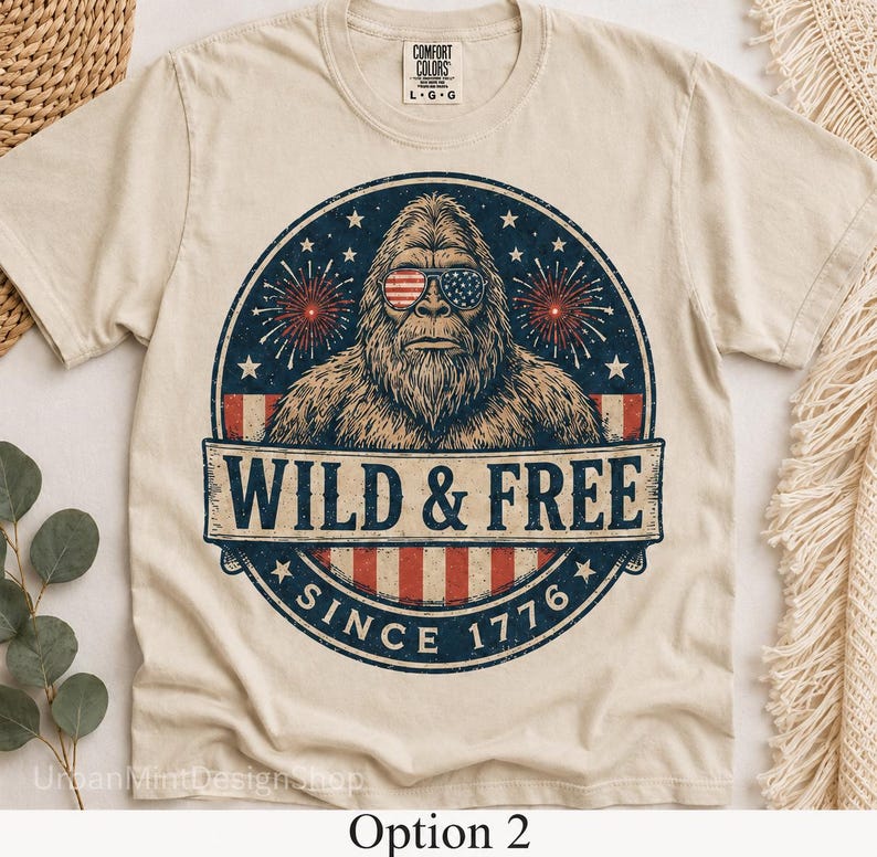 Puede incluir: Camiseta color crema con un dise&ntilde;o de Bigfoot con gafas de sol con la bandera estadounidense. El dise&ntilde;o incluye el texto "WILD & FREE SINCE 1776" en un emblema circular. Marca Comfort Colors.