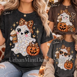 May include: A charcoal gray t-shirt featuring three graphic options of a ghost with a leopard print bow, holding a coffee cup and a jack-o'-lantern. The designs include sparkling accents, stars, and hearts. The text UrbanMintDesignShop is visible.