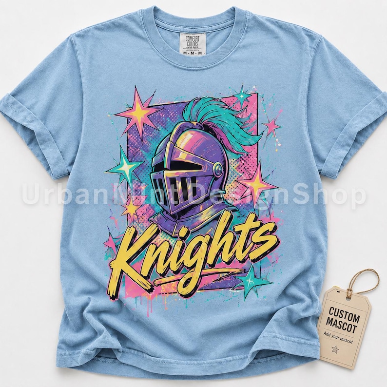 May include: A light blue t-shirt featuring a graphic of a purple knight helmet with a teal plume. The design includes yellow text that reads Knights, surrounded by pink and teal stars and a rectangular background pattern with paint splatters.