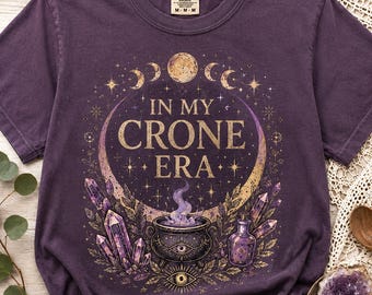 May include: A dark purple short-sleeve t-shirt featuring a graphic print with the text "IN MY CRONE ERA". The design includes a central cauldron with purple smoke, surrounded by crystals, stars, moon phases, and a small bottle, all in gold and purple tones.