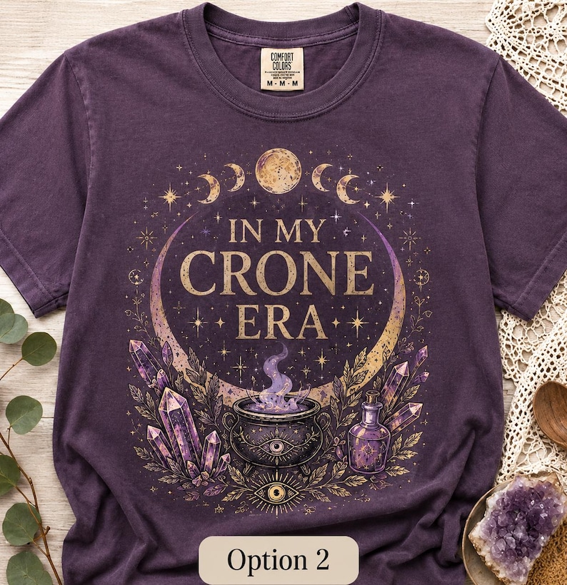 K&ouml;nnte beinhalten: Ein dunkelviolettes Kurzarm-T-Shirt mit einem Grafikdruck und dem Text "IN MY CRONE ERA". Das Design zeigt einen zentralen Kessel mit violettem Rauch, umgeben von Kristallen, Sternen, Mondphasen und einer kleinen Flasche in Gold- und Violettt&ouml;nen.