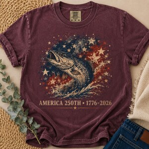 May include: A maroon short-sleeve t-shirt featuring a graphic of a jumping fish surrounded by white stars and a red, white, and blue distressed background. The text reads AMERICA 250TH 1776-2026. The shirt has a crew neckline and a vintage wash finish.