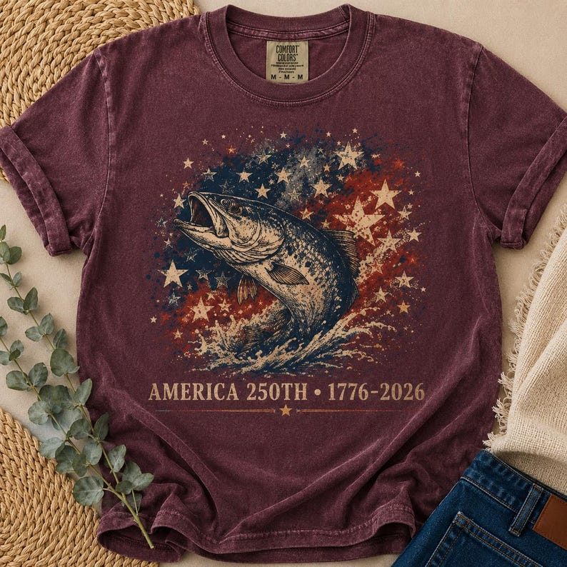 May include: A maroon short-sleeve t-shirt featuring a graphic of a jumping fish surrounded by white stars and a red, white, and blue distressed background. The text reads AMERICA 250TH 1776-2026. The shirt has a crew neckline and a vintage wash finish.