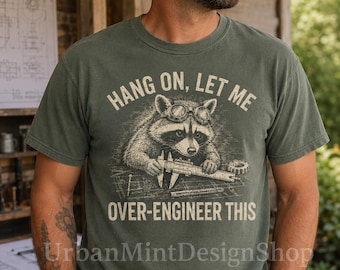 Puede incluir: Una camiseta verde salvia de cuello redondo con un gráfico de un mapache con gafas protectoras usando un calibre sobre un plano. El texto HANG ON, LET ME OVER-ENGINEER THIS aparece en color crema. Incluye un engranaje y un clavo.