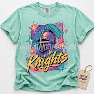 May include: A mint green short-sleeve t-shirt featuring a graphic of a purple knight helmet with a teal plume. The design includes yellow cursive text that reads Knights, surrounded by pink and teal stars and a rectangular background pattern.