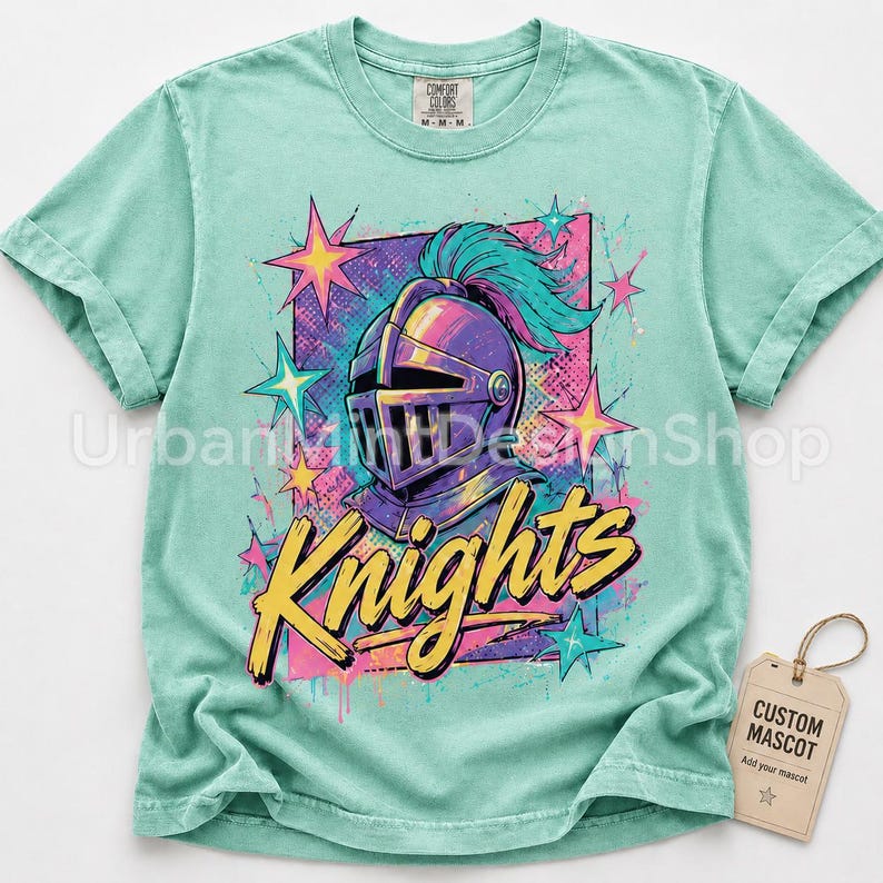 May include: A mint green short-sleeve t-shirt featuring a graphic of a purple knight helmet with a teal plume. The design includes yellow cursive text that reads Knights, surrounded by pink and teal stars and a rectangular background pattern.